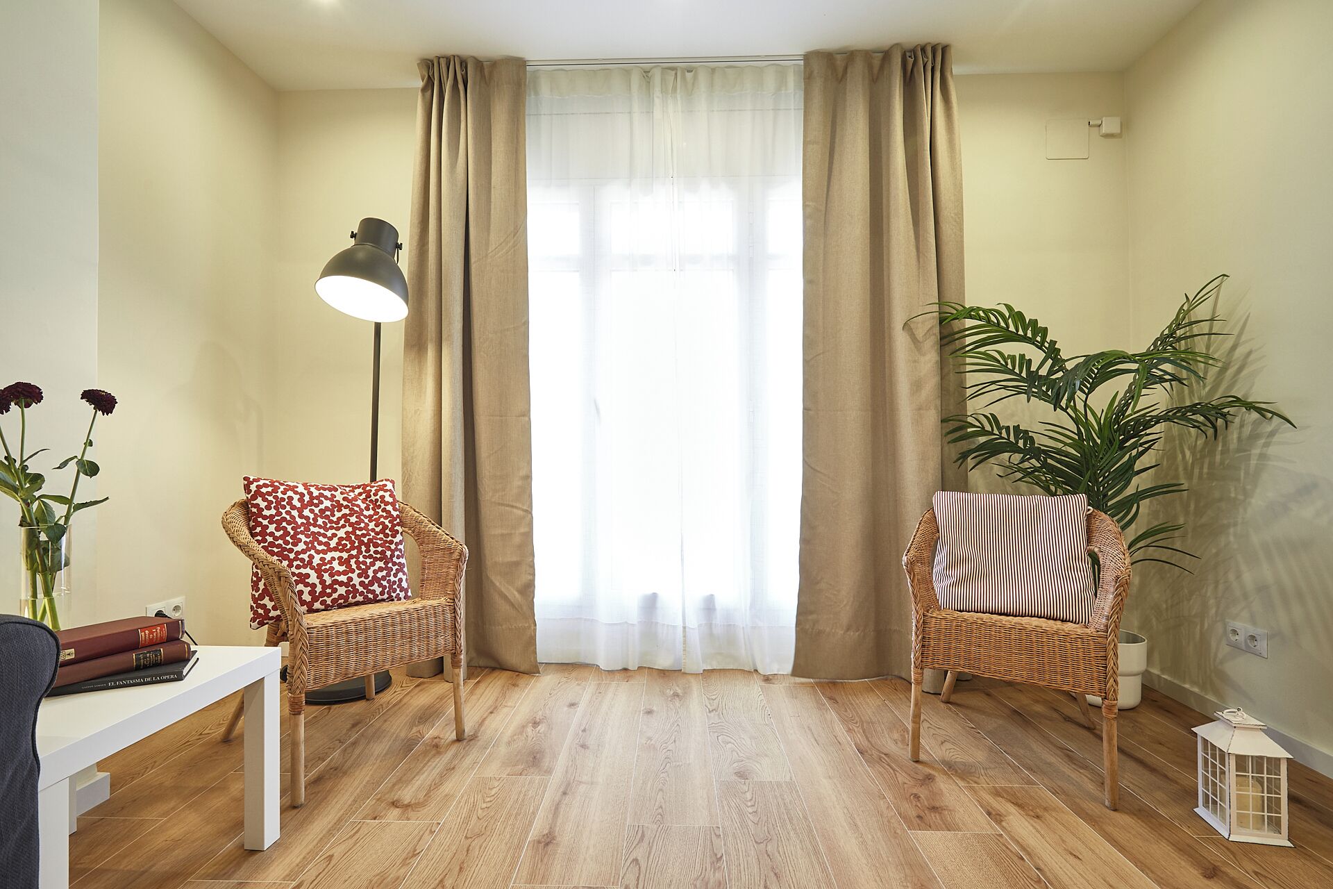 A living room with two wicker chairs, a floor lamp, a potted plant, and a white table with books.