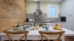 Una cocina con pared de ladrillo, encimera blanca y ventana. Hay dos sillas de madera, una mesa con artículos para el desayuno y un microondas.