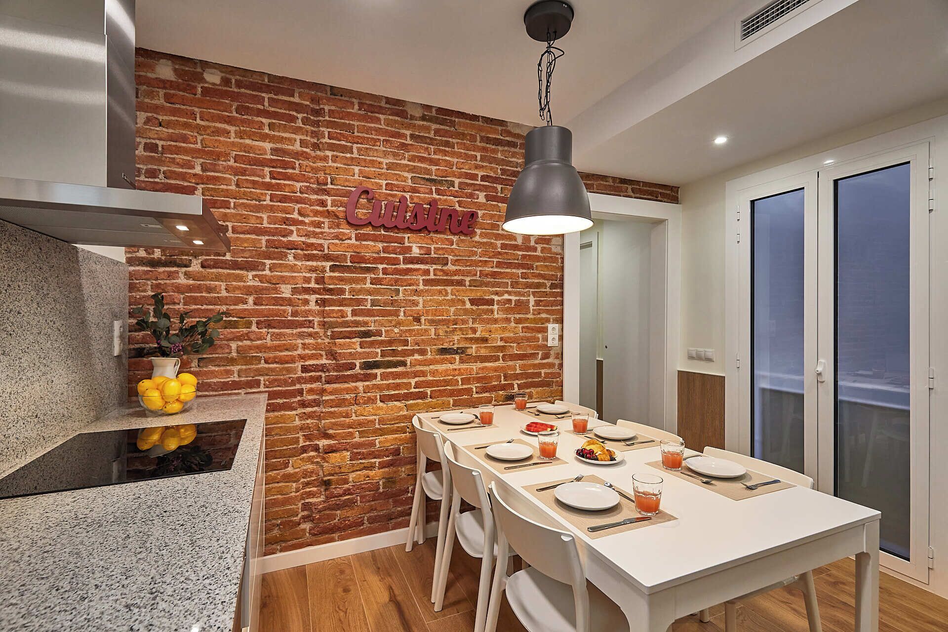 A modern kitchen with a brick wall, a dining area with a white table set for four, and a hanging light fixture.