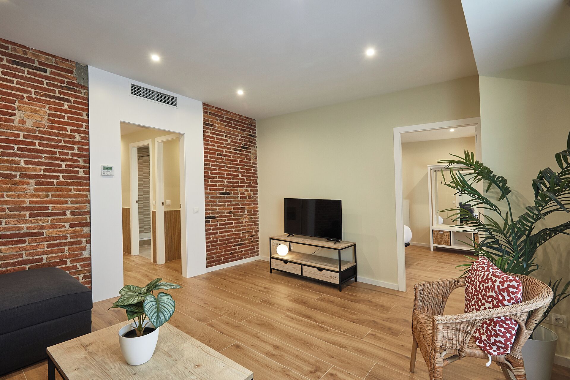 Una sala moderna con una pared de ladrillo decorativa, un televisor de pantalla plana sobre un soporte de madera, una silla de mimbre y una planta en maceta.