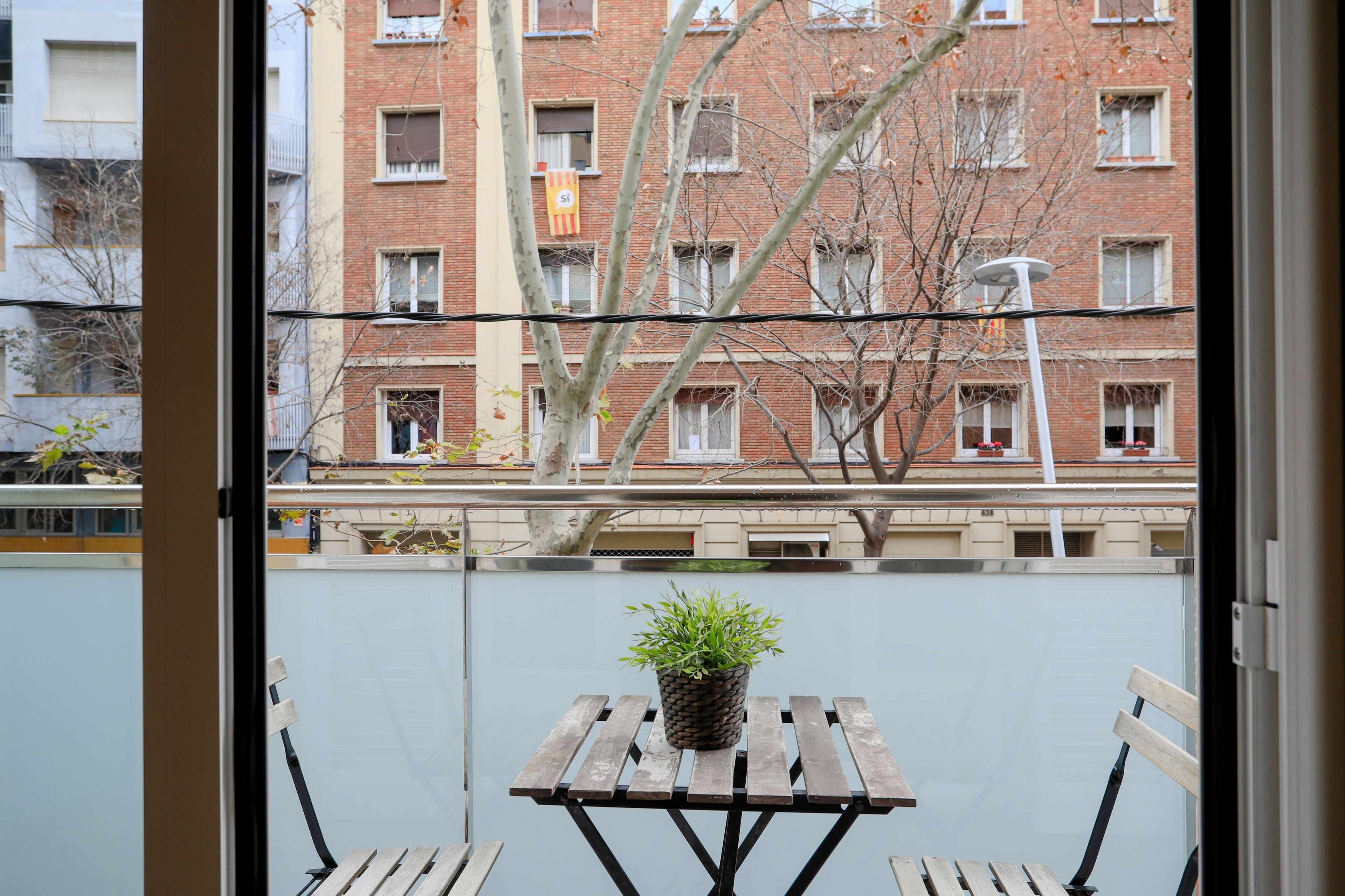 Un balcon avec une table et des chaises en bois, une plante en pot et une vue sur un immeuble en briques doté de nombreuses fenêtres et de balcons.