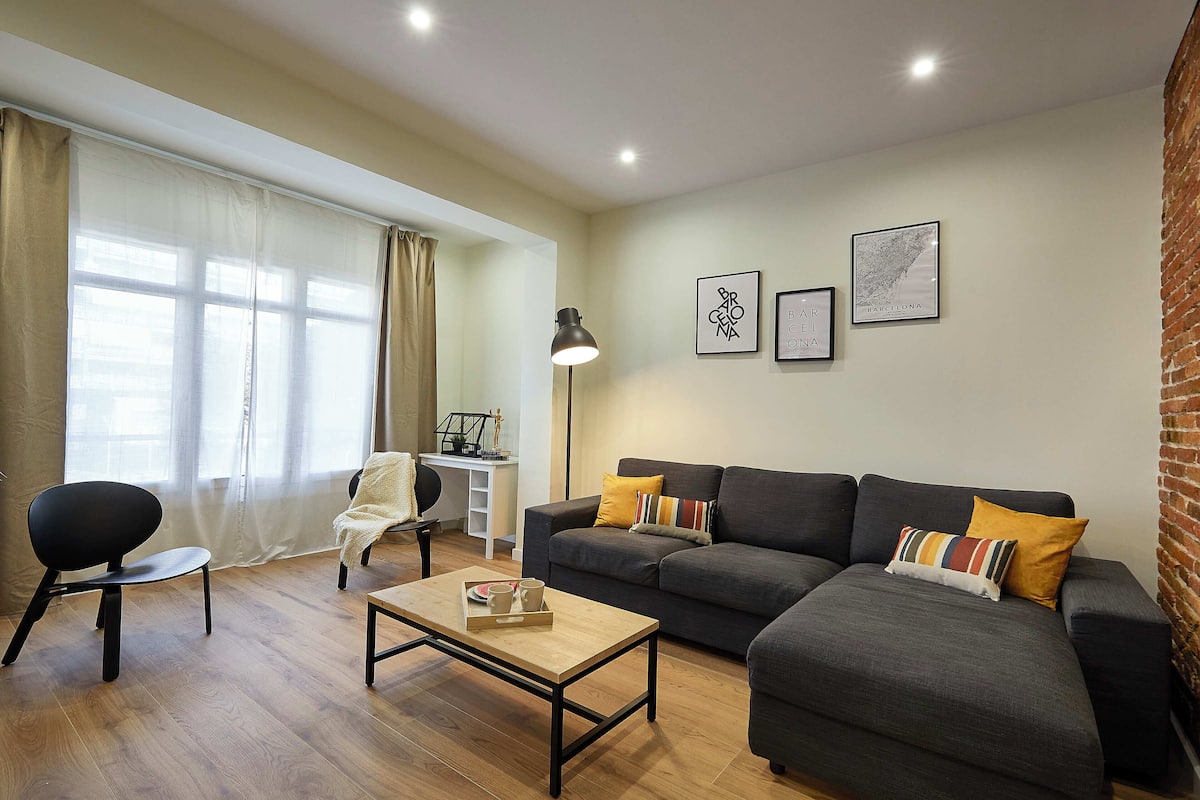 A modern living room with a grey sofa, a wooden coffee table, and a black chair. There are two framed pictures on the wall and a lamp on a side table.