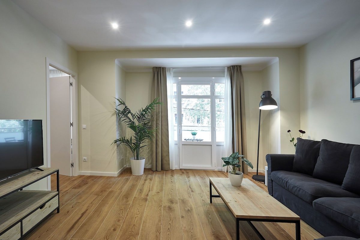 A modern living room with a wooden floor, a dark sofa, a TV on a wooden stand, and two potted plants.