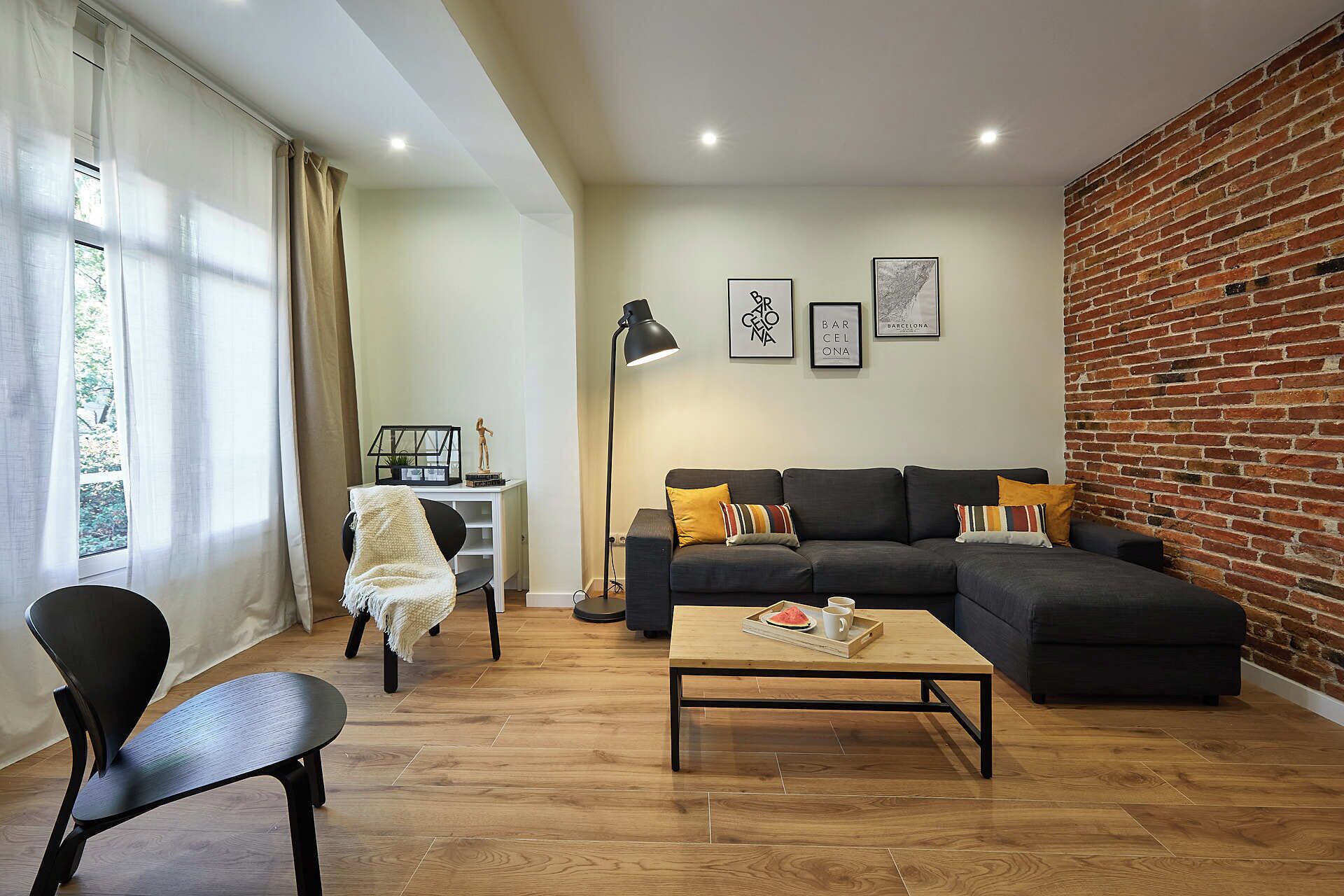 A living room with a sectional sofa, a coffee table, a black chair, a desk with a lamp, and a brick wall.