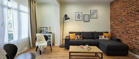 A living room with a sectional sofa, a coffee table, a black chair, a desk with a lamp, and a brick wall.