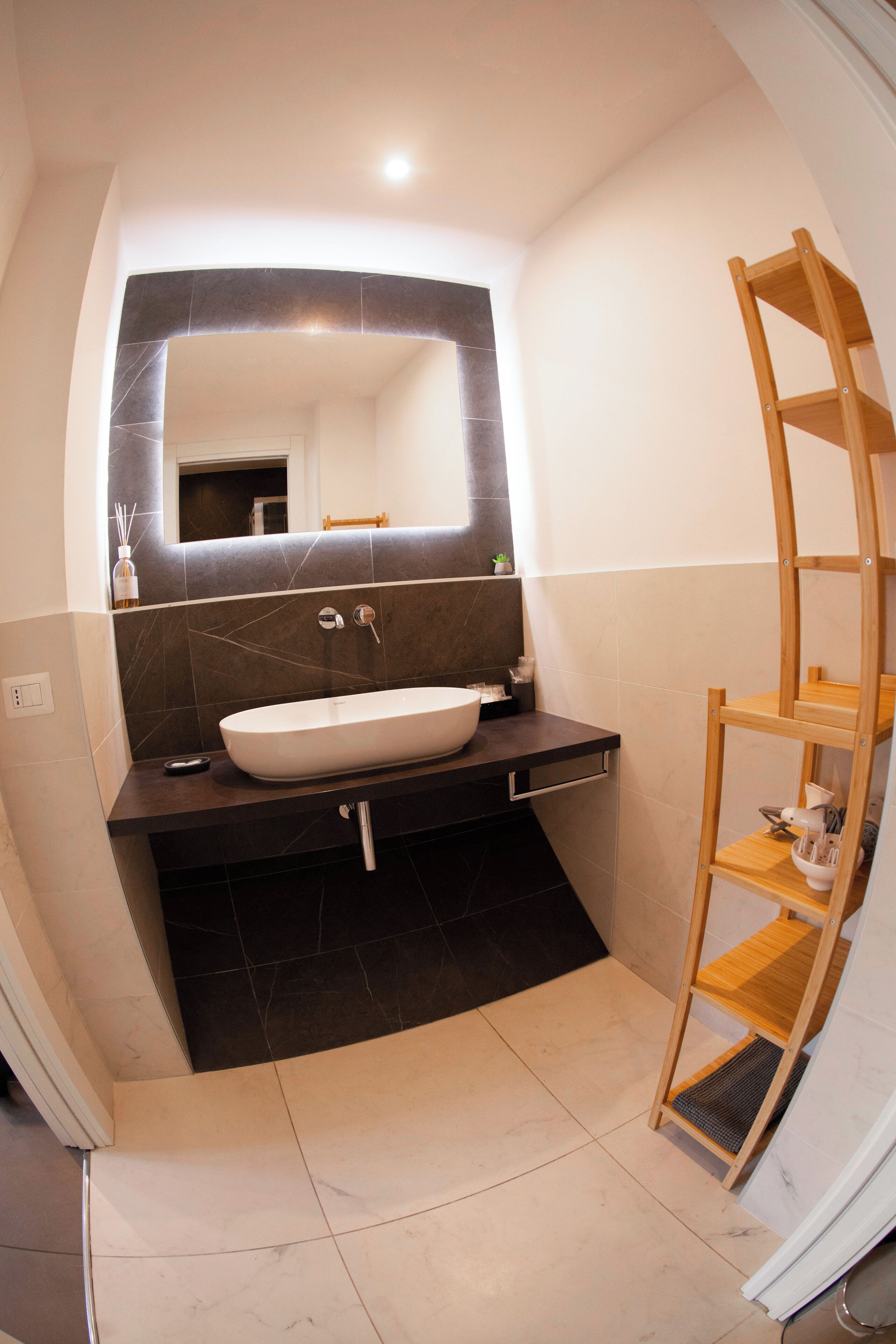 Een moderne badkamer met een grote spiegel, een witte wastafel, donkere tegels en een houten plank.