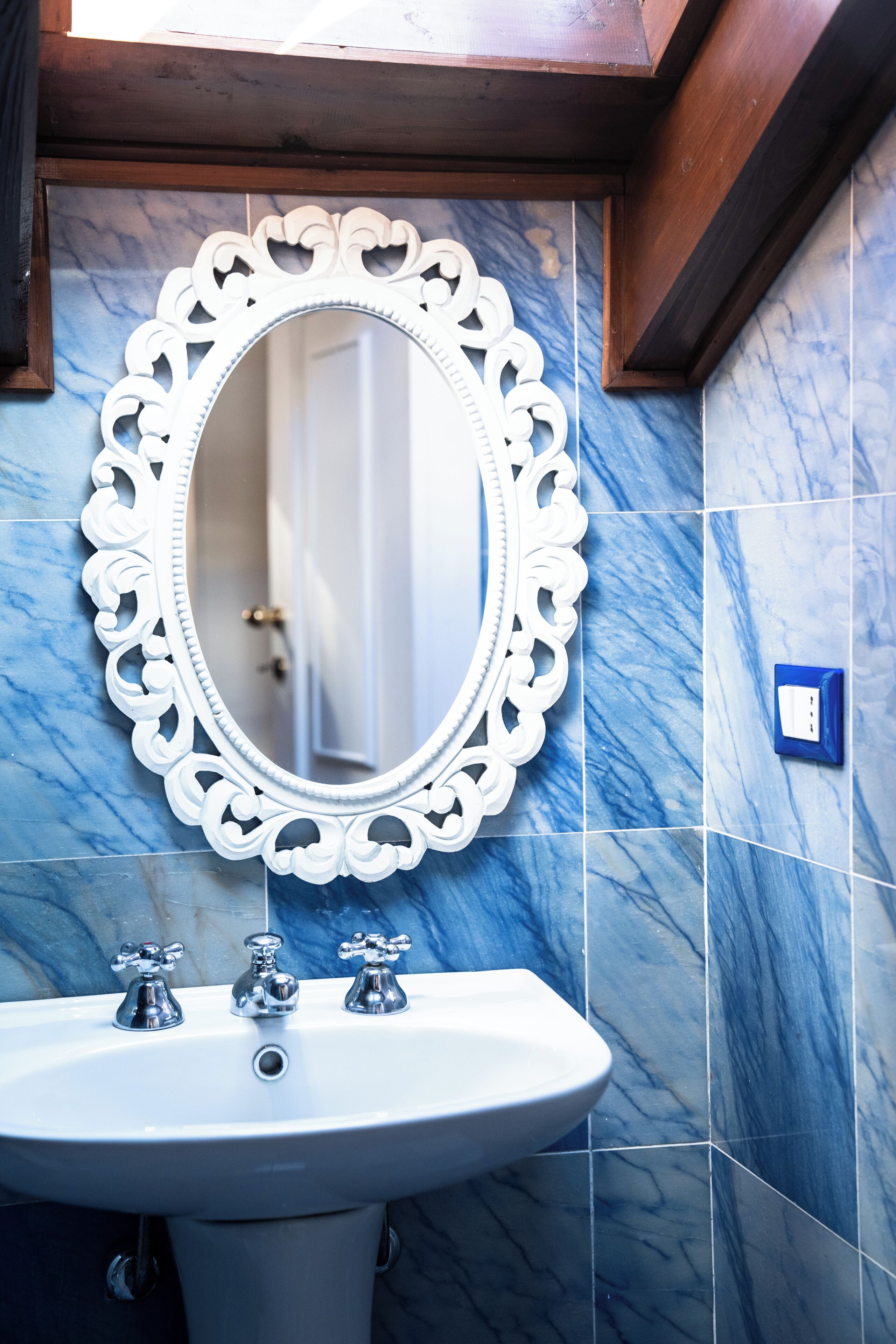 Ein Badezimmer mit einem weißen, verzierten Spiegel, einem weißen Waschbecken mit drei Wasserhähnen und blauen Marmorfliesen.