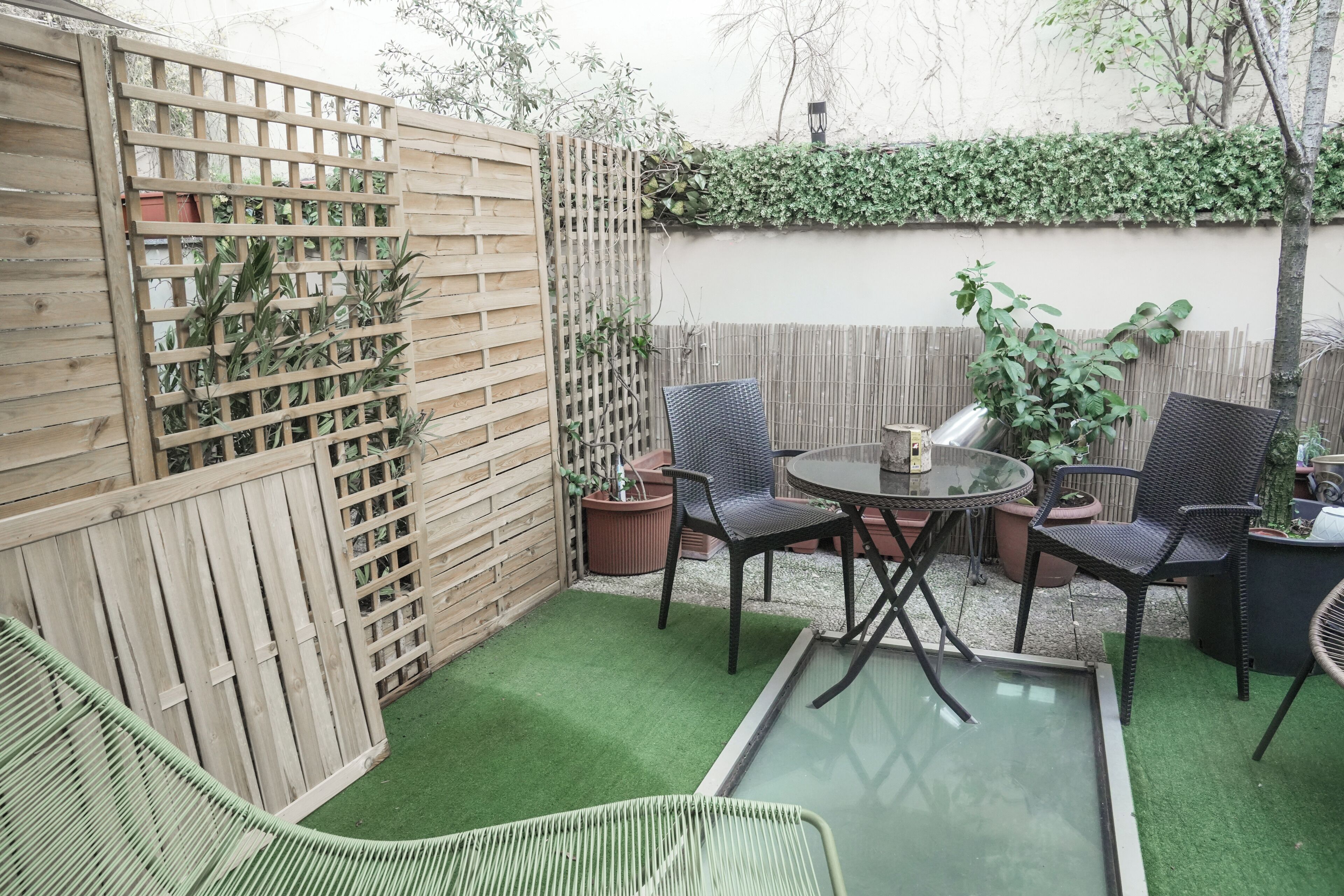 A patio with a wooden fence, two chairs, a small table, and a potted plant.
