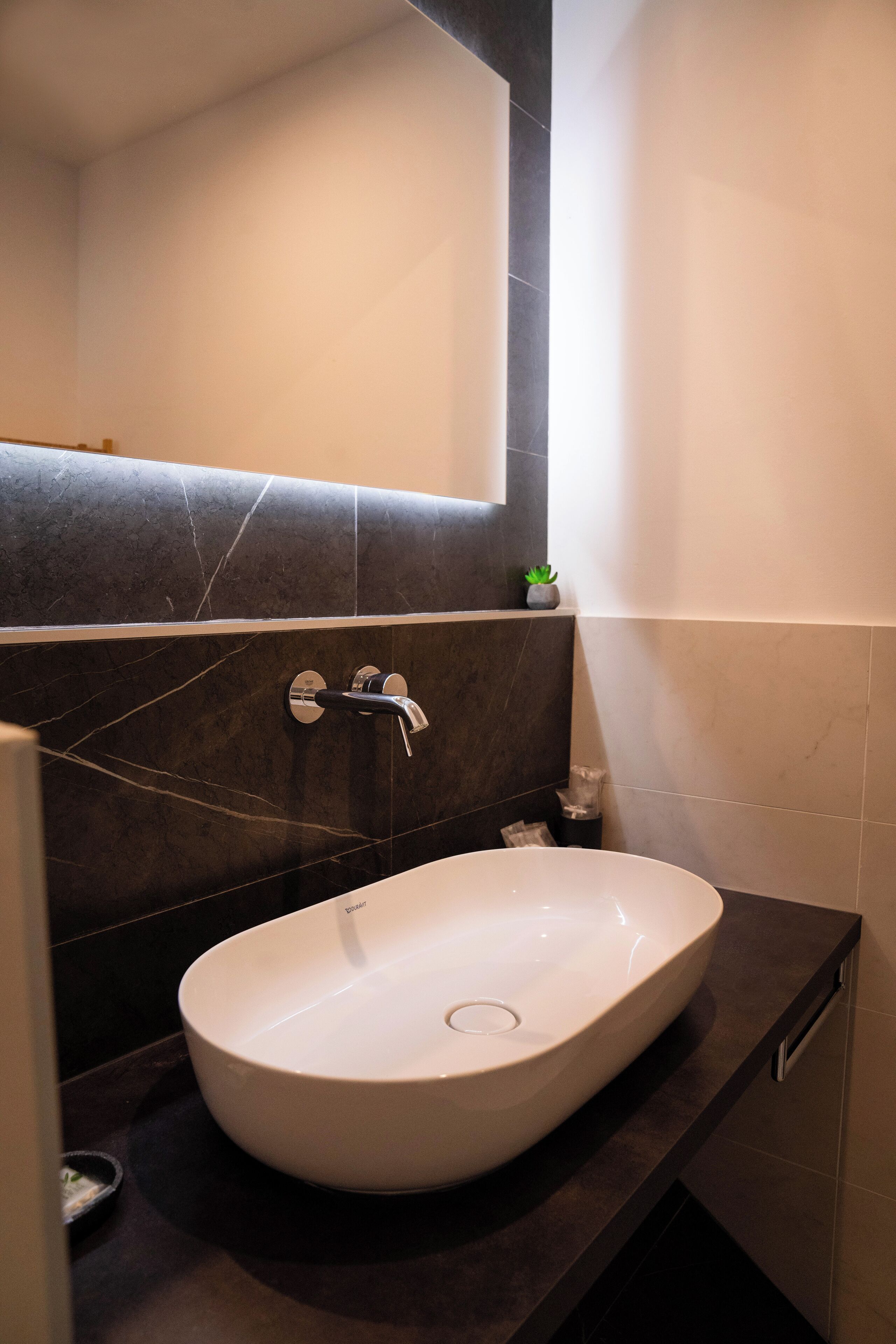 A modern bathroom with a large oval sink, dark marble walls, and a small potted plant on the counter.