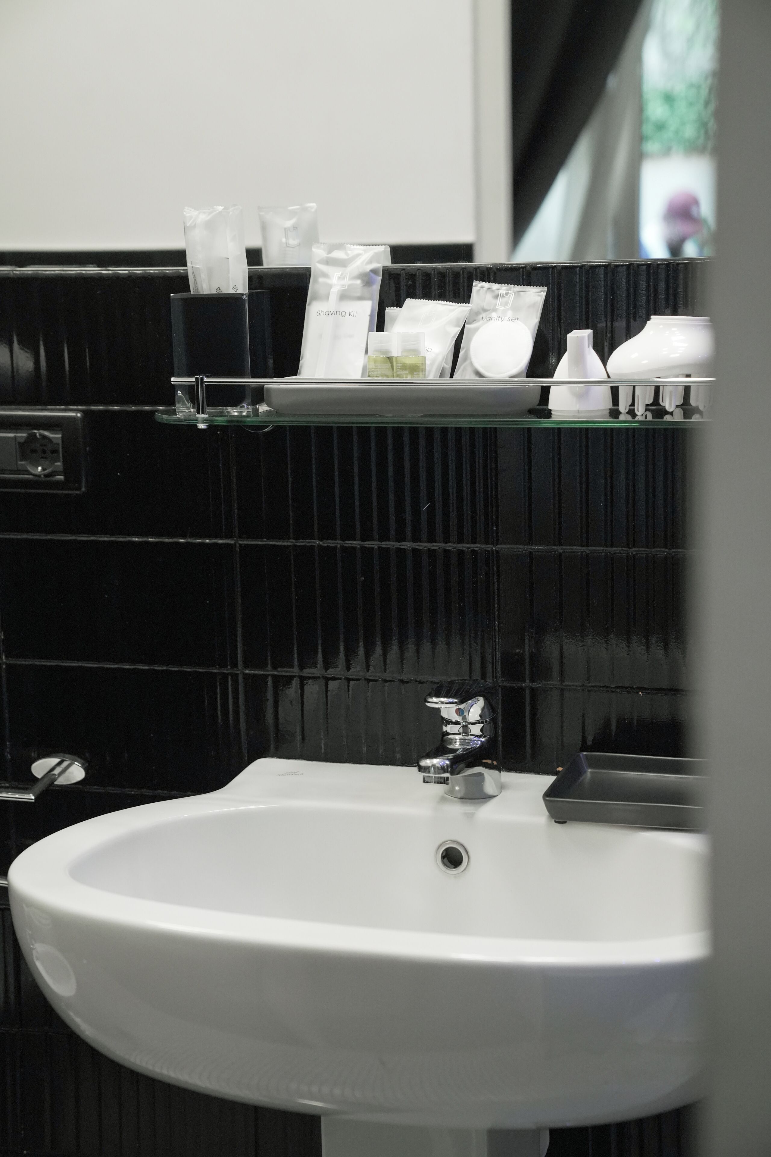 A bathroom with a white sink, black tiled walls, and a shelf with toiletries.
