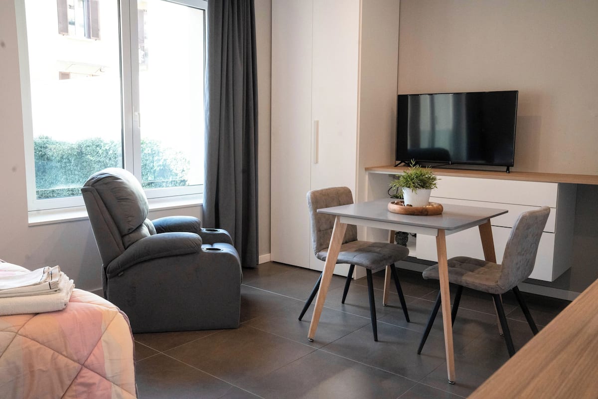 A modern hotel room with a bed, a seating area with a table and chairs, a flat-screen TV mounted on the wall, and a window with curtains.