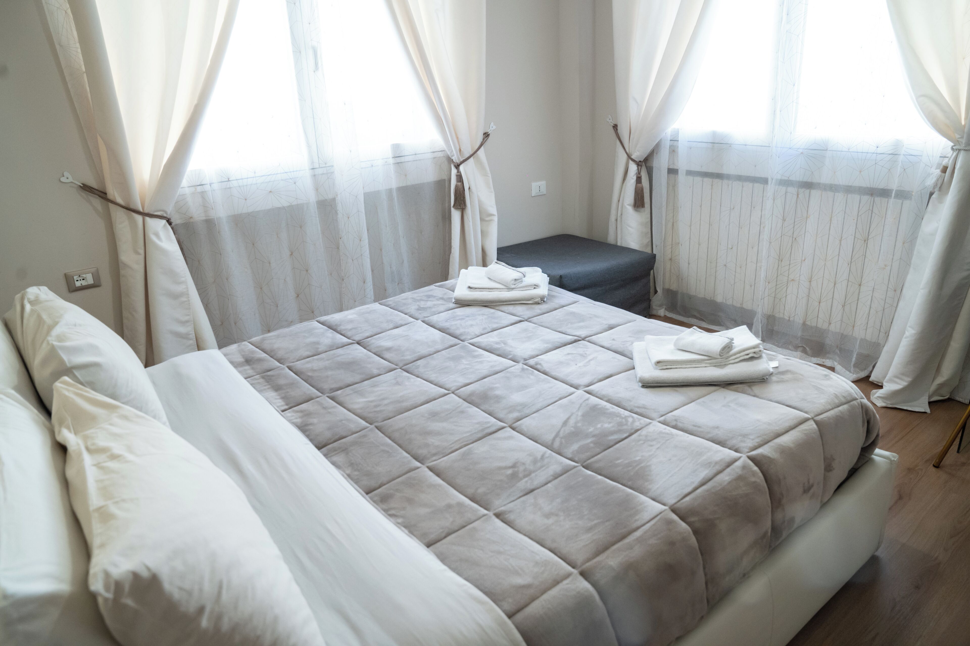 A neatly made bed with a grey quilt, a small bedside table with a folded towel, and sheer curtains on a window.
