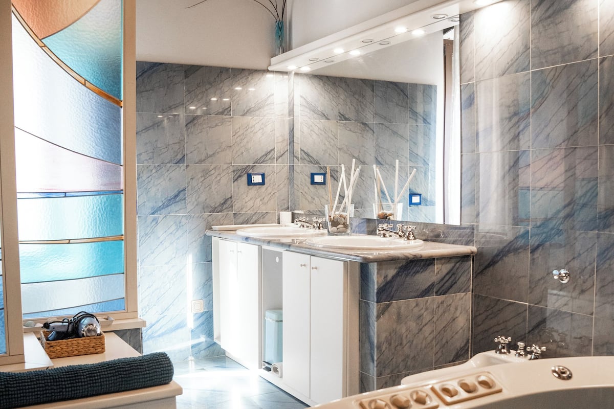 A modern bathroom with a marble wall, a large bathtub, a vanity with a mirror, and a window with a blue view.