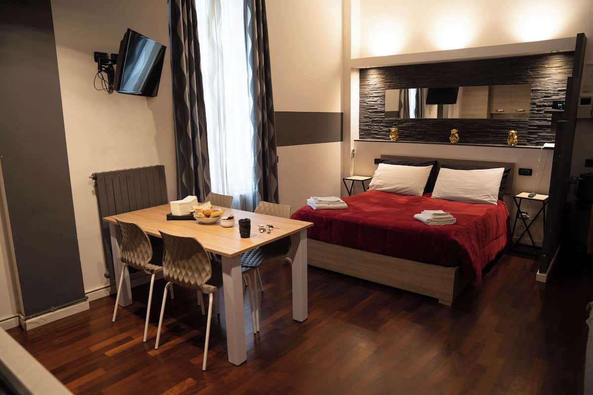 A hotel room with a bed, a dining table with chairs, a TV mounted on the wall, and a window with curtains.