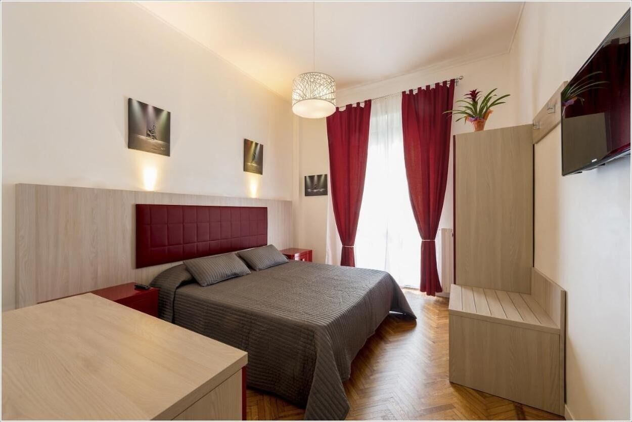 A modern hotel room with a large bed, a wooden nightstand, a desk, a TV, and a potted plant.
