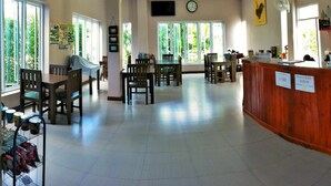 A spacious dining area with multiple tables and chairs, large windows with greenery outside, a reception desk, and a clock on the wall. - Blue House Sukhothai (Sukhothai)