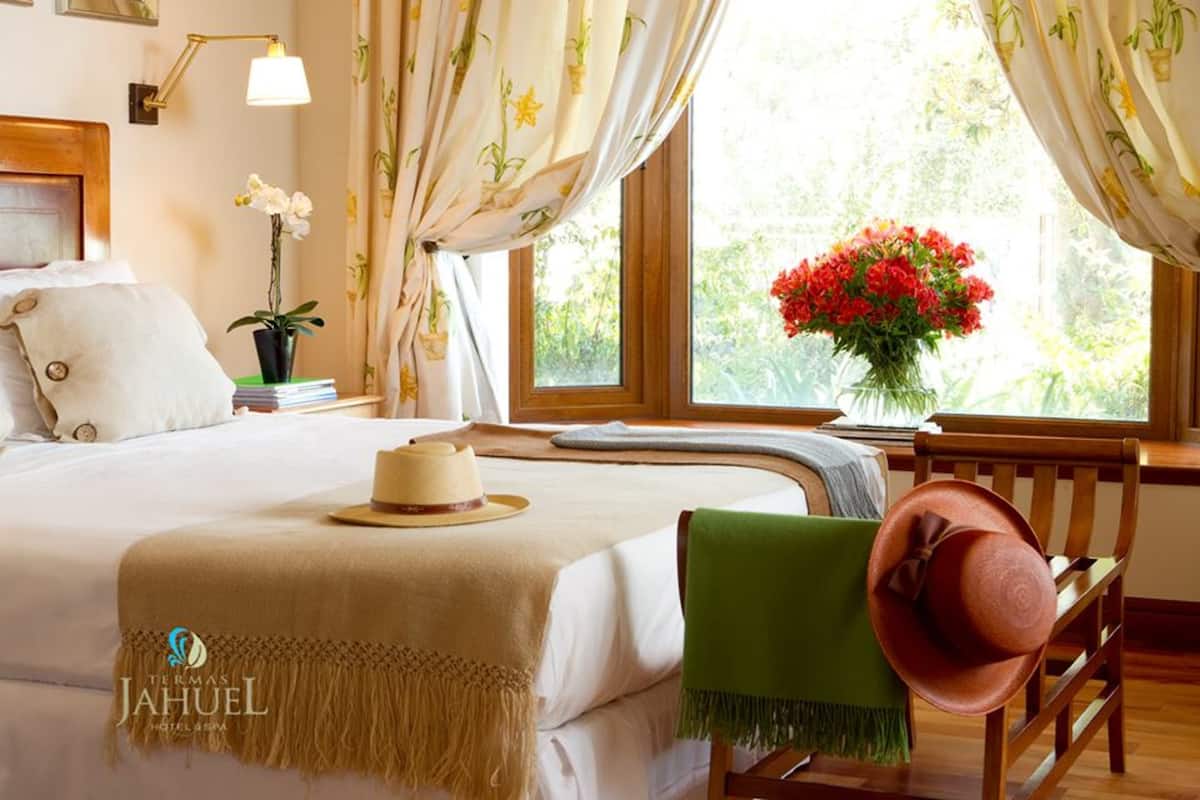 A hotel room with a bed, a nightstand with a lamp, a window with curtains, a vase of flowers, and a hat on a chair.