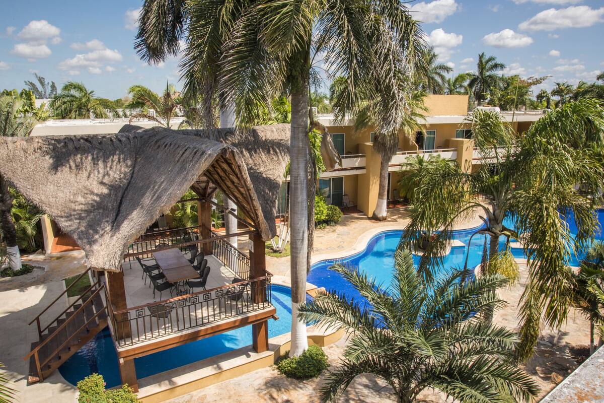 A resort with a pool, palm trees, and a thatched-roof lounge area.