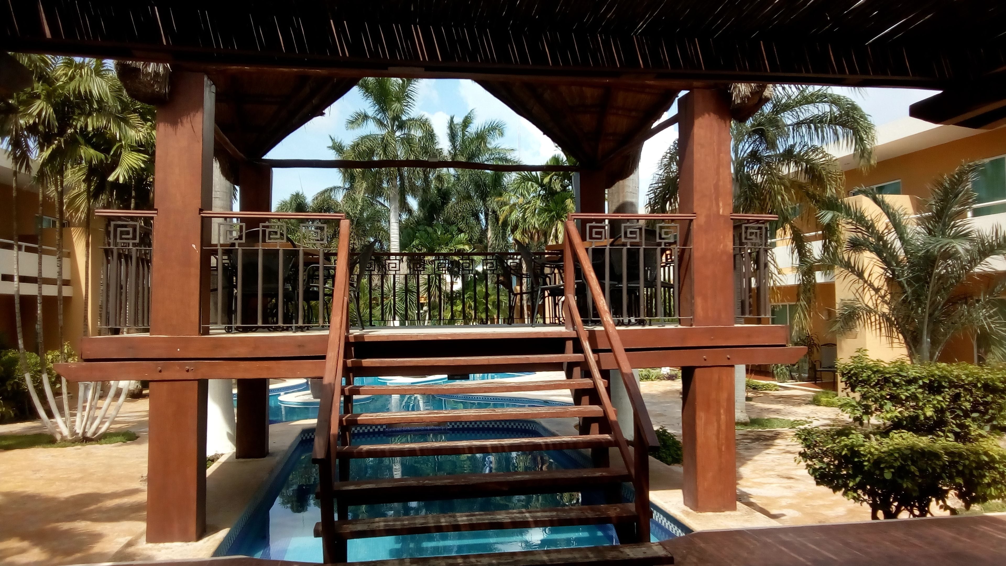 Zona de piscina con una escalera de madera que lleva a una terraza cubierta, rodeada de palmeras y un edificio con una verja geométrica.