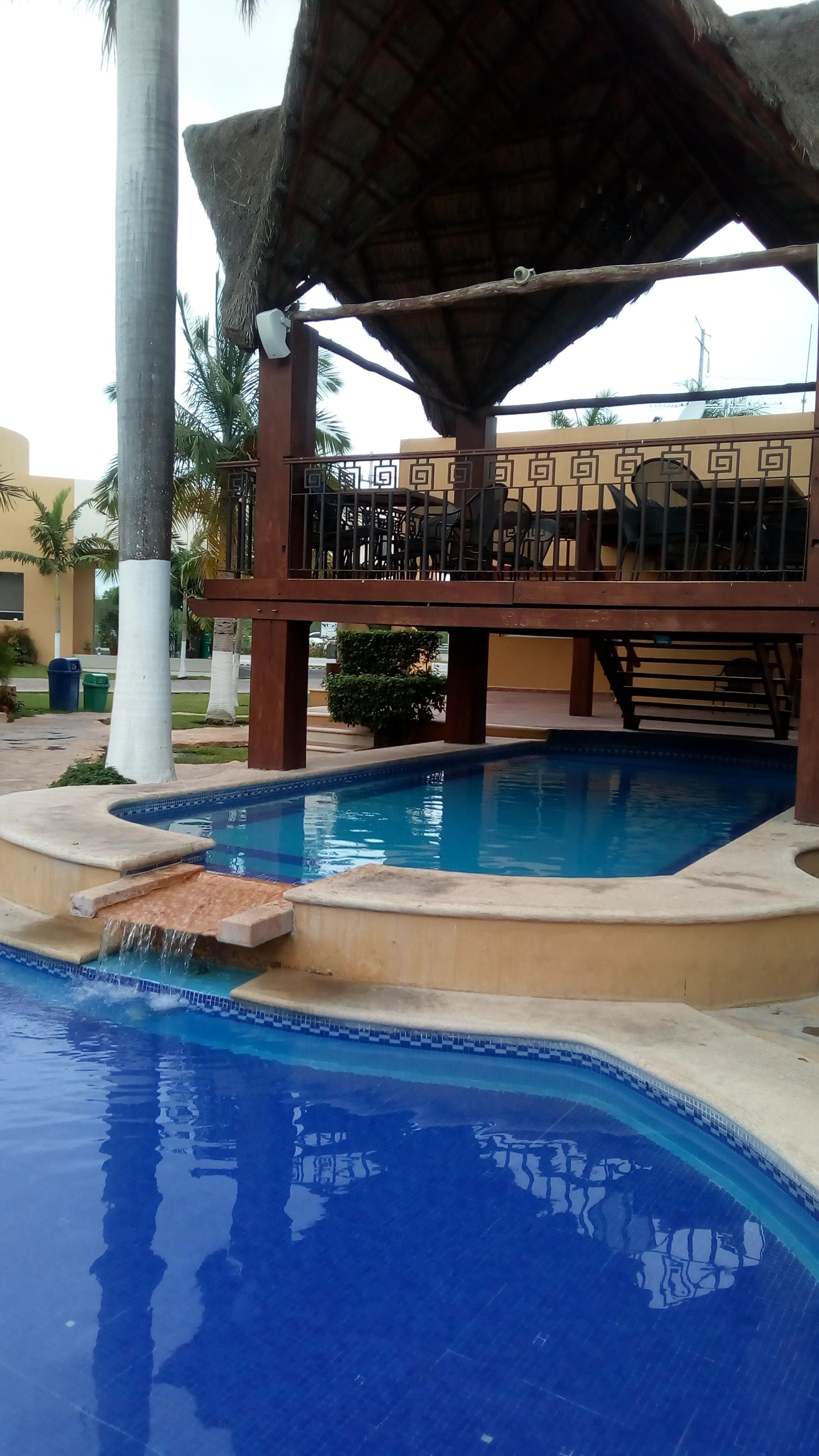 Una piscina con fuente, zona de estar techada y palmeras al fondo.