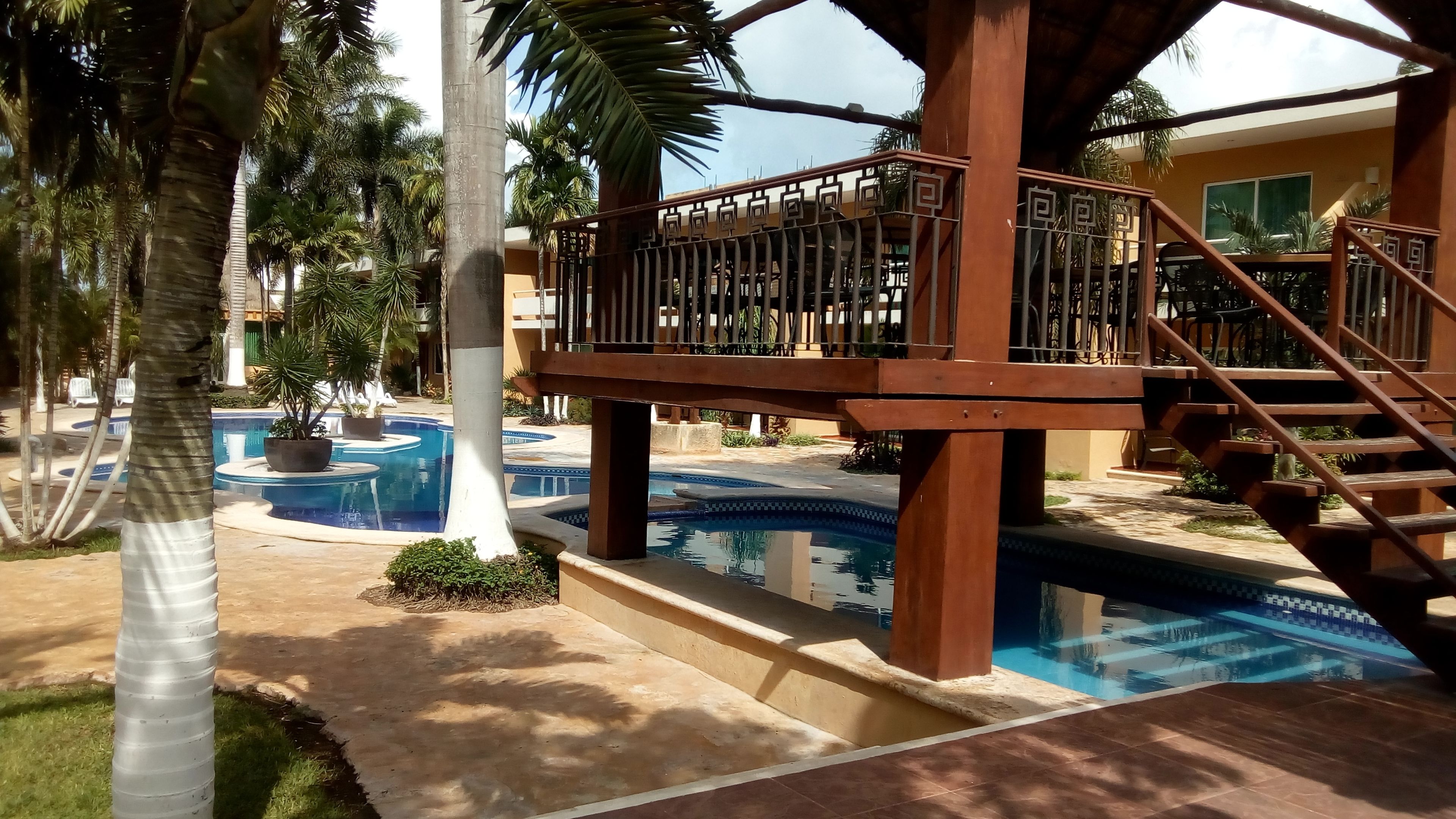 Ein Poolbereich mit Holzterrasse und Treppe, umgeben von Palmen und einem Gebäude mit gelber Fassade.