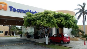 A hotel entrance with a large sign, a red car parked in front, and a tree in front of the entrance. - TecnoHotel Mérida Norte (Mérida)