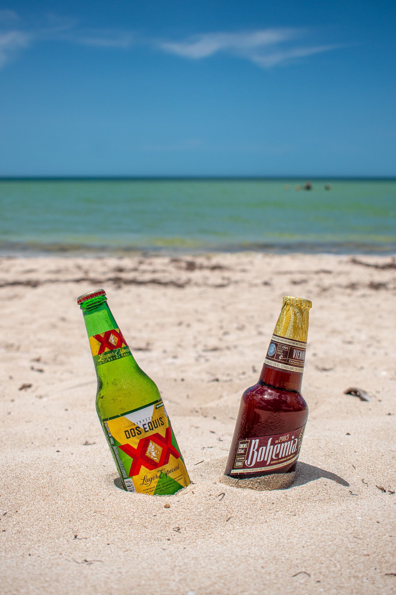 Dos botellas de cerveza sobre una playa arenosa, con un cielo azul despejado y el océano al fondo.