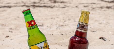 Dos botellas de cerveza sobre una playa arenosa, con un cielo azul despejado y el océano al fondo.
