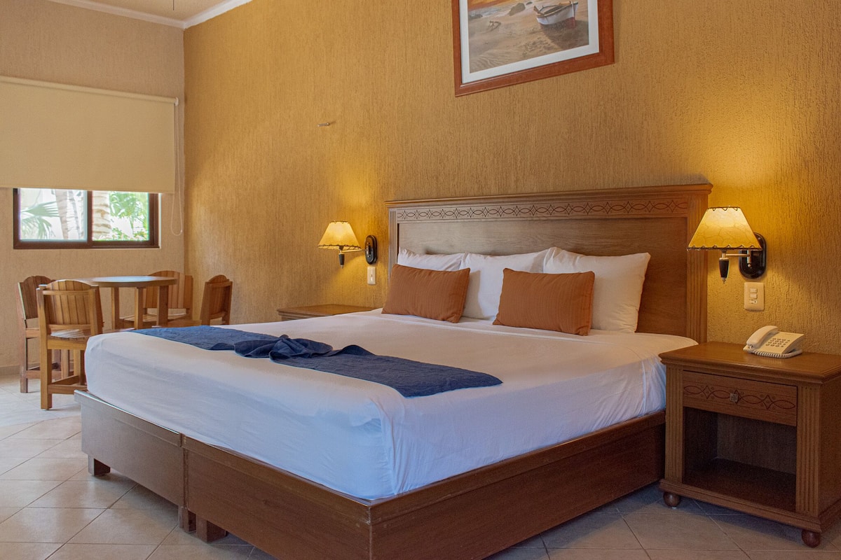 A hotel room with a large bed, two bedside lamps, a nightstand with a phone, a wooden chair, and a small table.