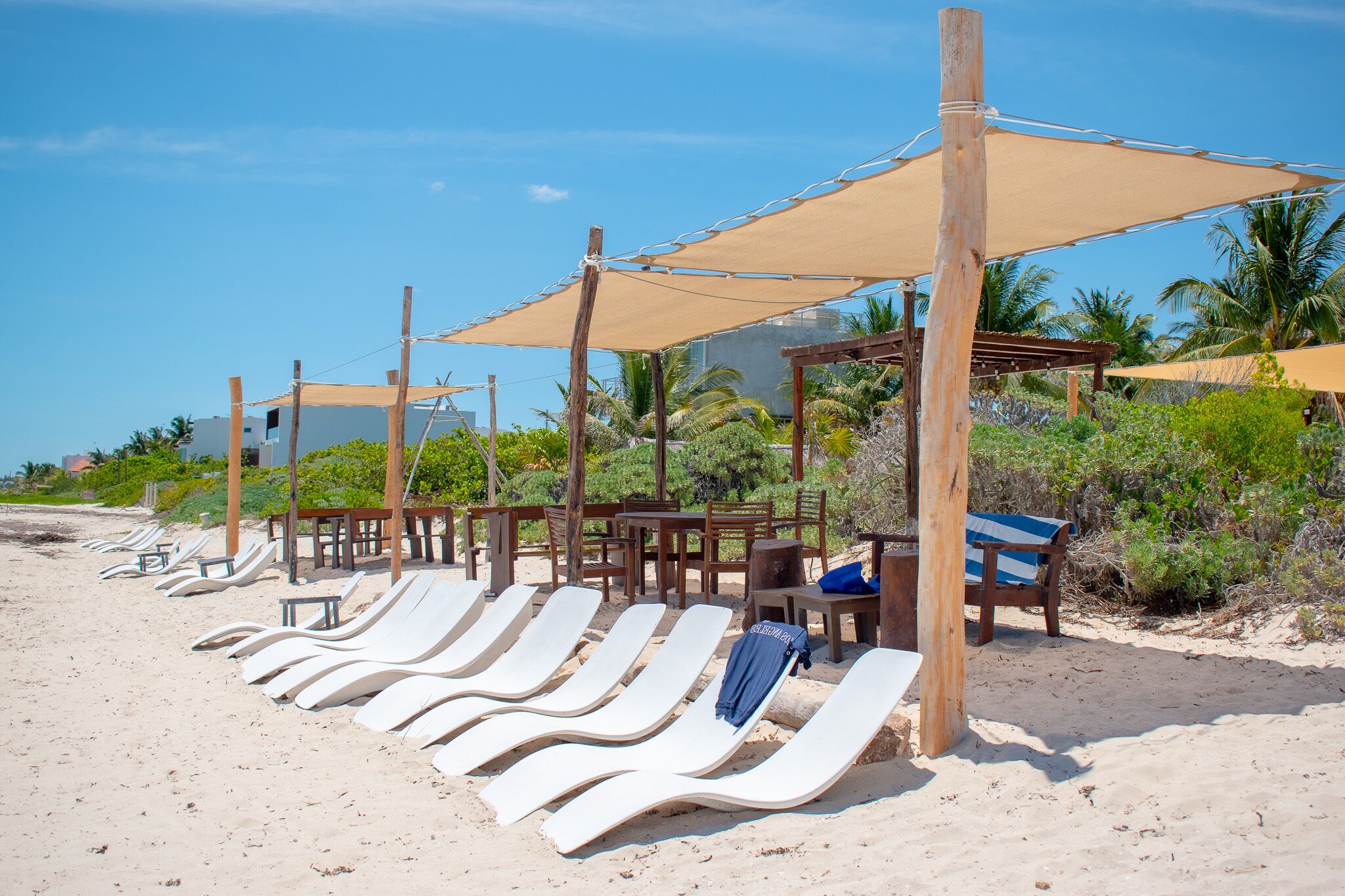 A beachside lounge area with sun loungers and wooden tables under shaded canopies.