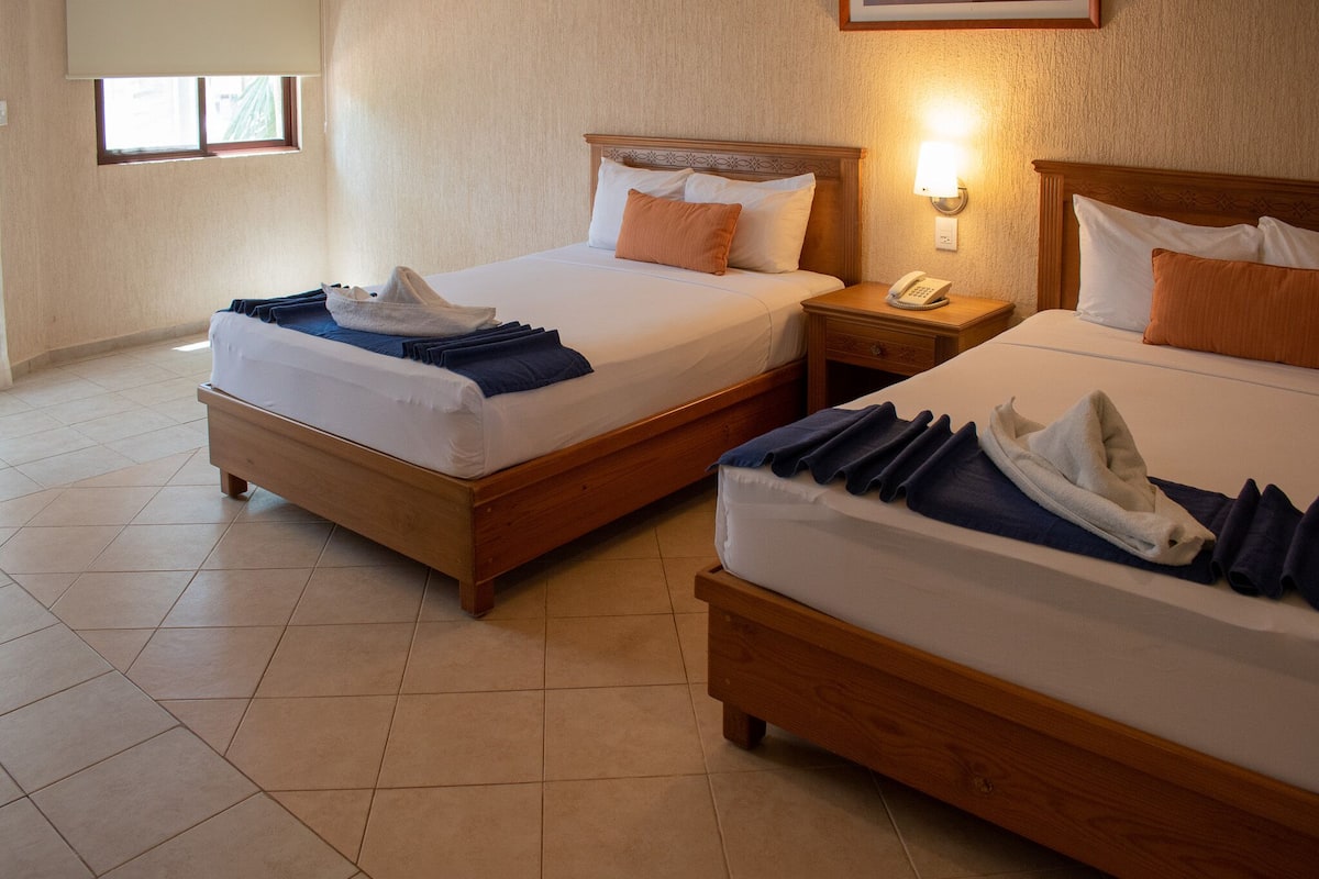 Two wooden beds with white linens and blue towels, a bedside table with a lamp, and a window with blinds.
