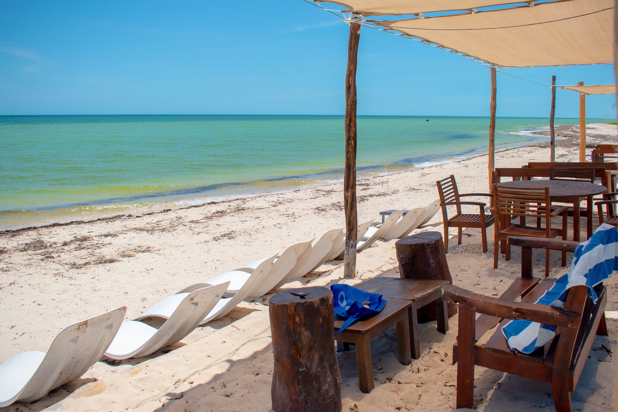 A beachside seating area with wooden tables and chairs, a covered area, and white lounge chairs lined up along the sand.