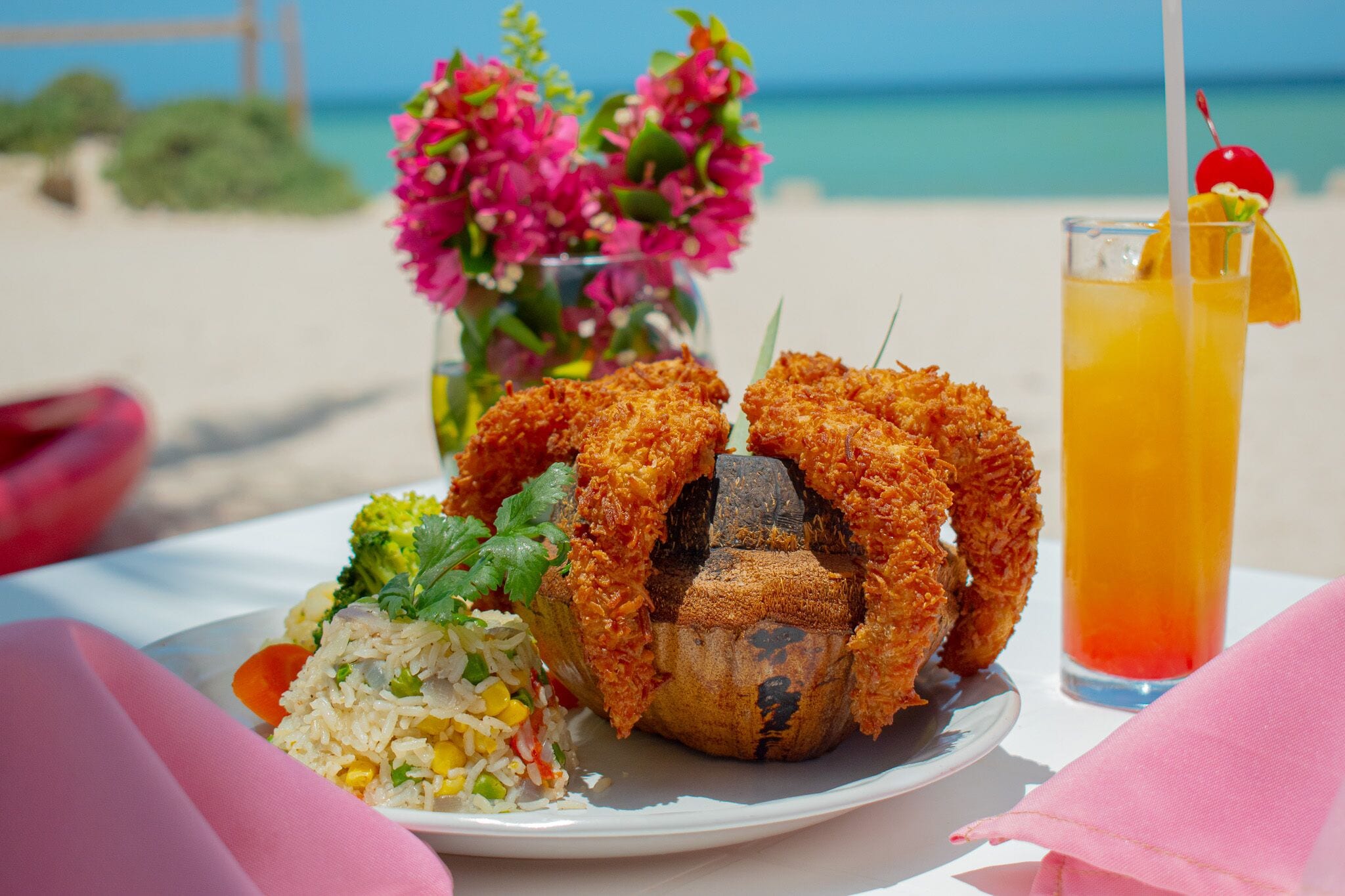 A plate of fried food, a drink, and a vase of flowers on a table by the beach.