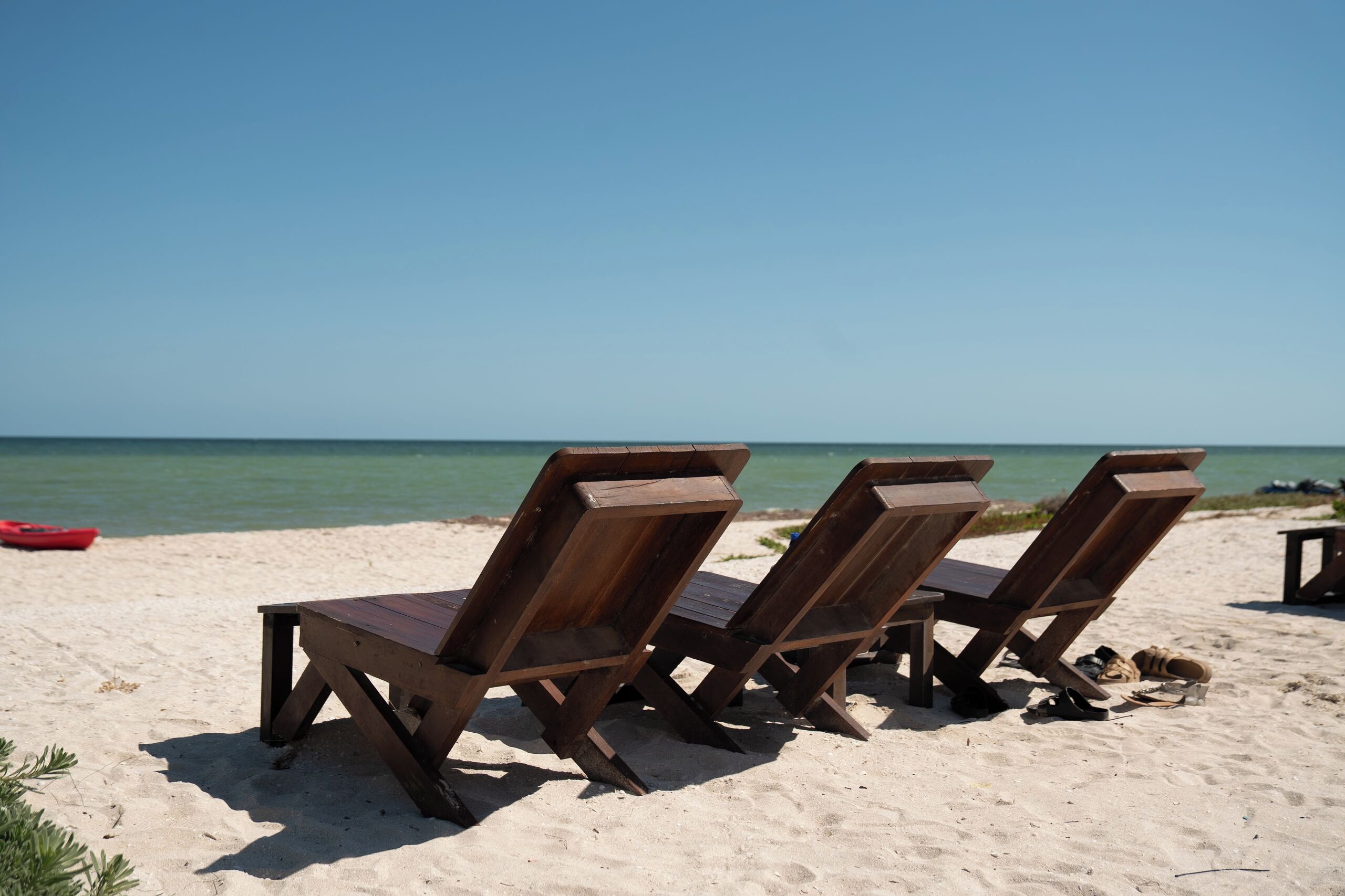 Three wooden beach chairs on a sandy beach with a clear blue sky and calm sea in the background.