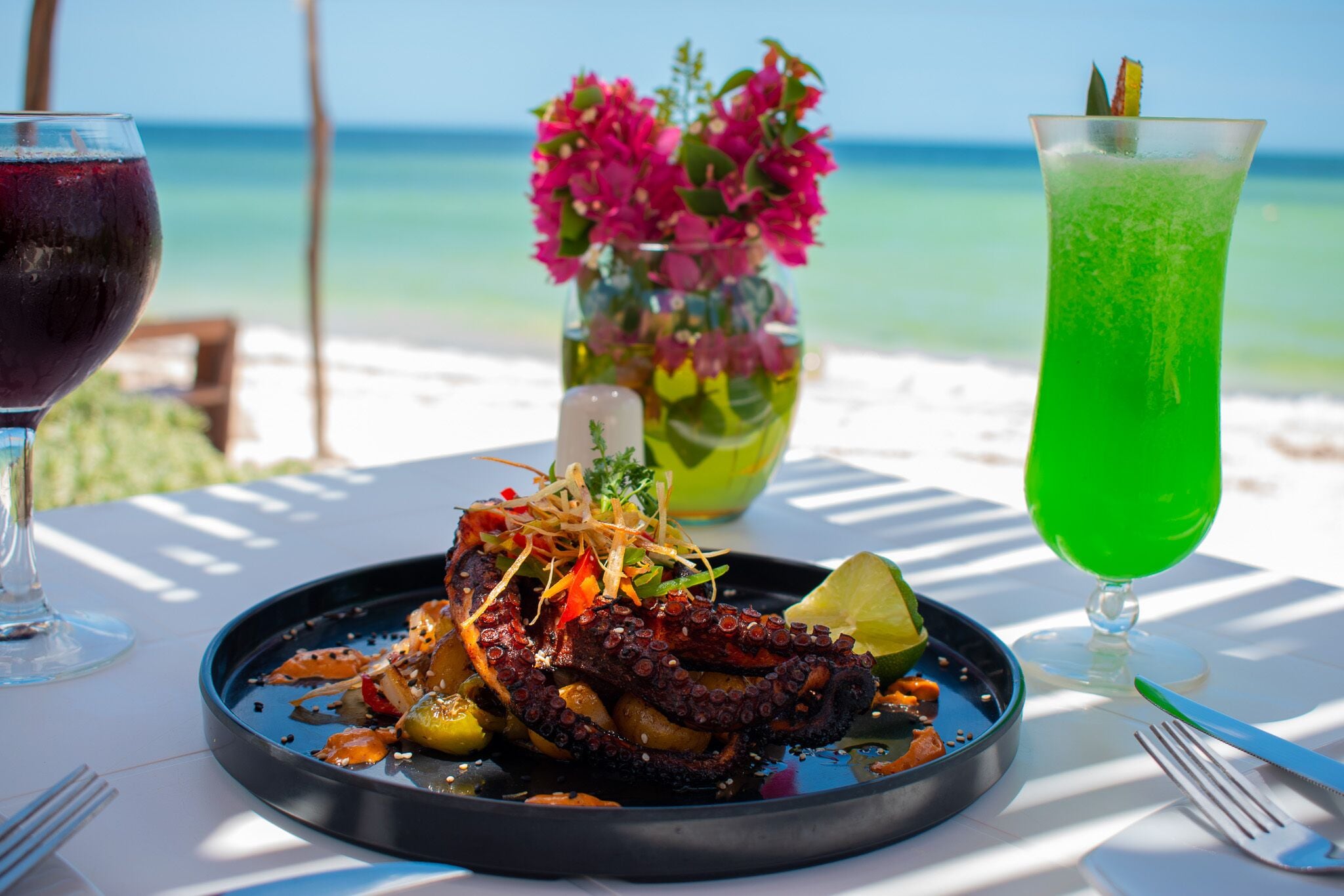 Un plato con pulpo y verduras, una copa de vino tinto y una bebida verde sobre una mesa con vistas a la playa.