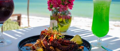 Un plato con pulpo y verduras, una copa de vino tinto y una bebida verde sobre una mesa con vistas a la playa.
