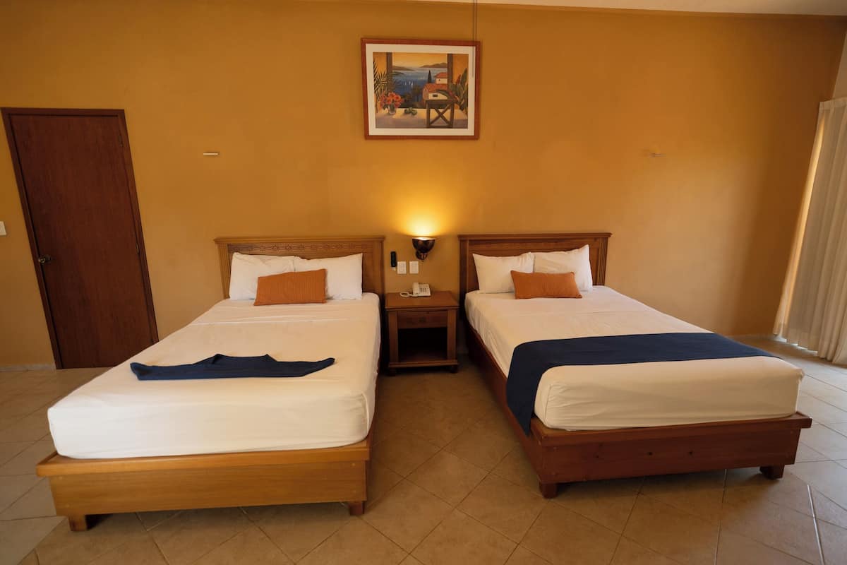 A hotel room with two single beds, a nightstand with a lamp, a wooden door, and a painting on the wall.