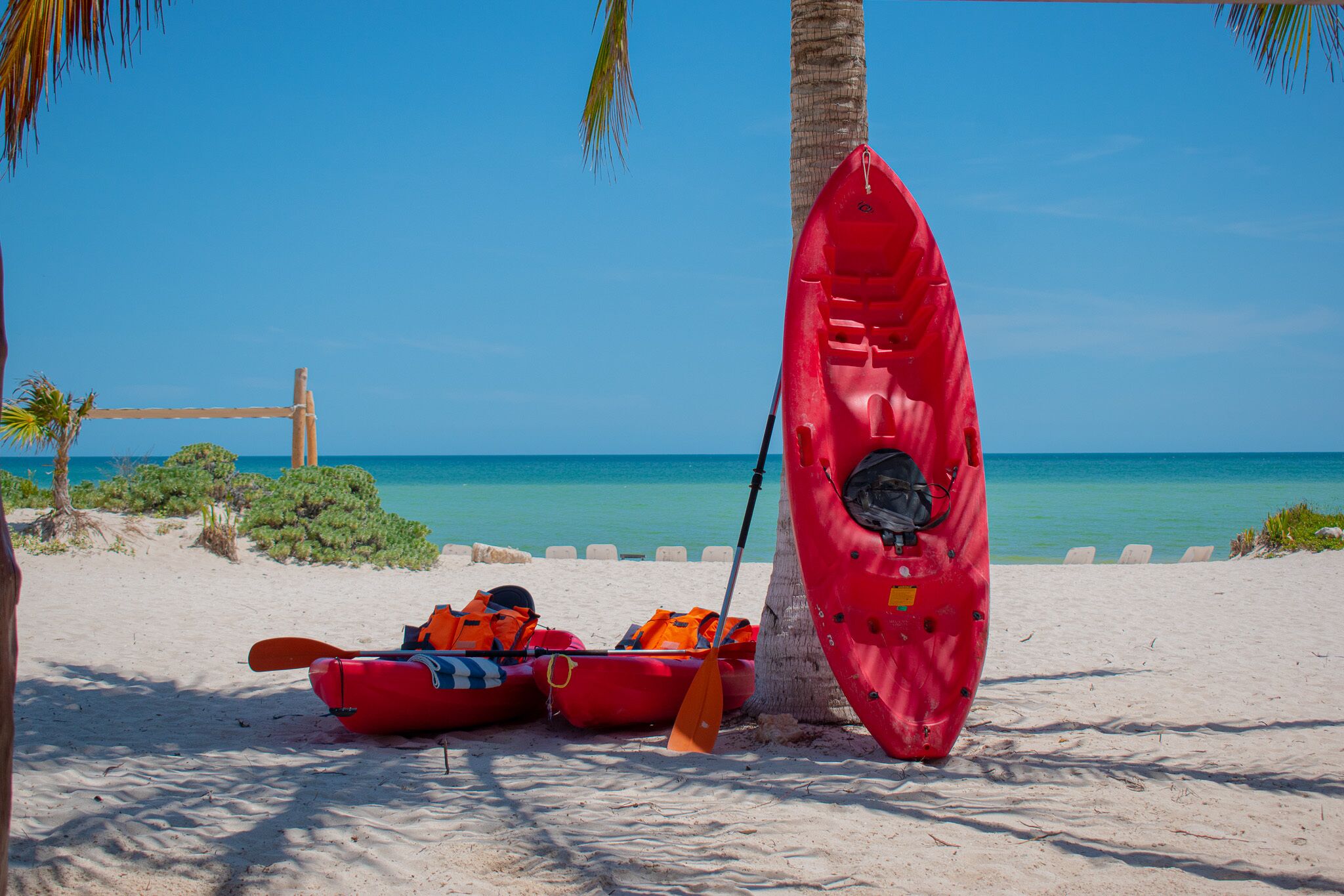 Un kayak rojo y una canoa roja sobre una playa arenosa, con un cielo azul despejado y el océano al fondo.