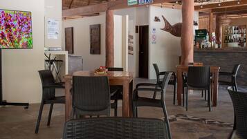Interior de restaurante com mesas e cadeiras de madeira, área de bar e uma grande tela de televisão na parede.