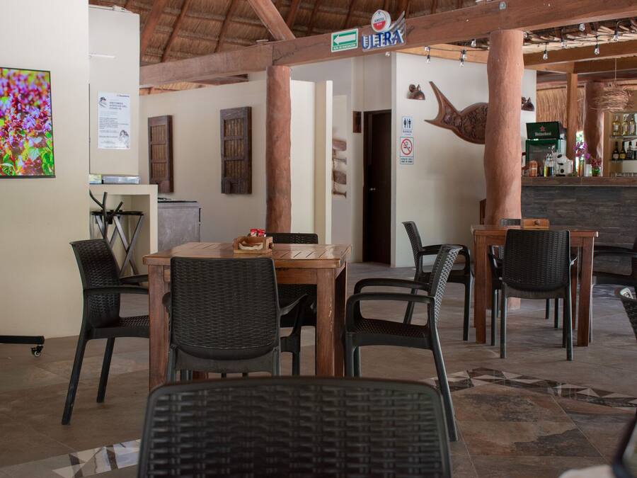 Interior de restaurante com mesas e cadeiras de madeira, área de bar e uma grande tela de televisão na parede.