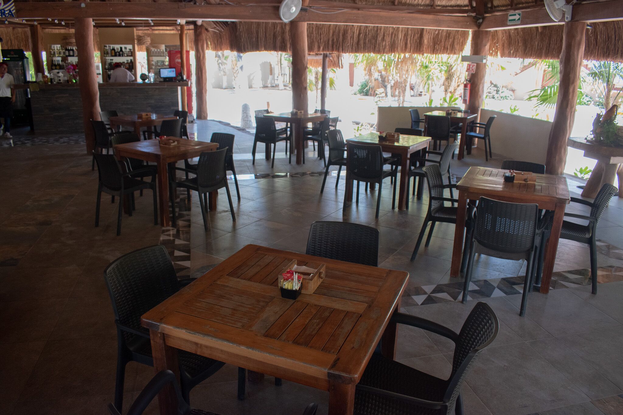 An outdoor restaurant with wooden tables and chairs, a bar area, and a view of the surrounding greenery.