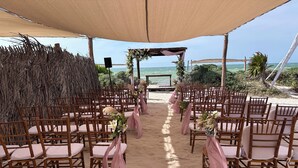 Un salón de bodas al aire libre con filas de sillas de madera y suelo de arena, dispuesto bajo una marquesina con vista al océano.