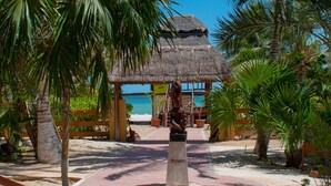 Un resort frente a la playa con una cabaña de techo de paja, palmeras y un cielo azul despejado.