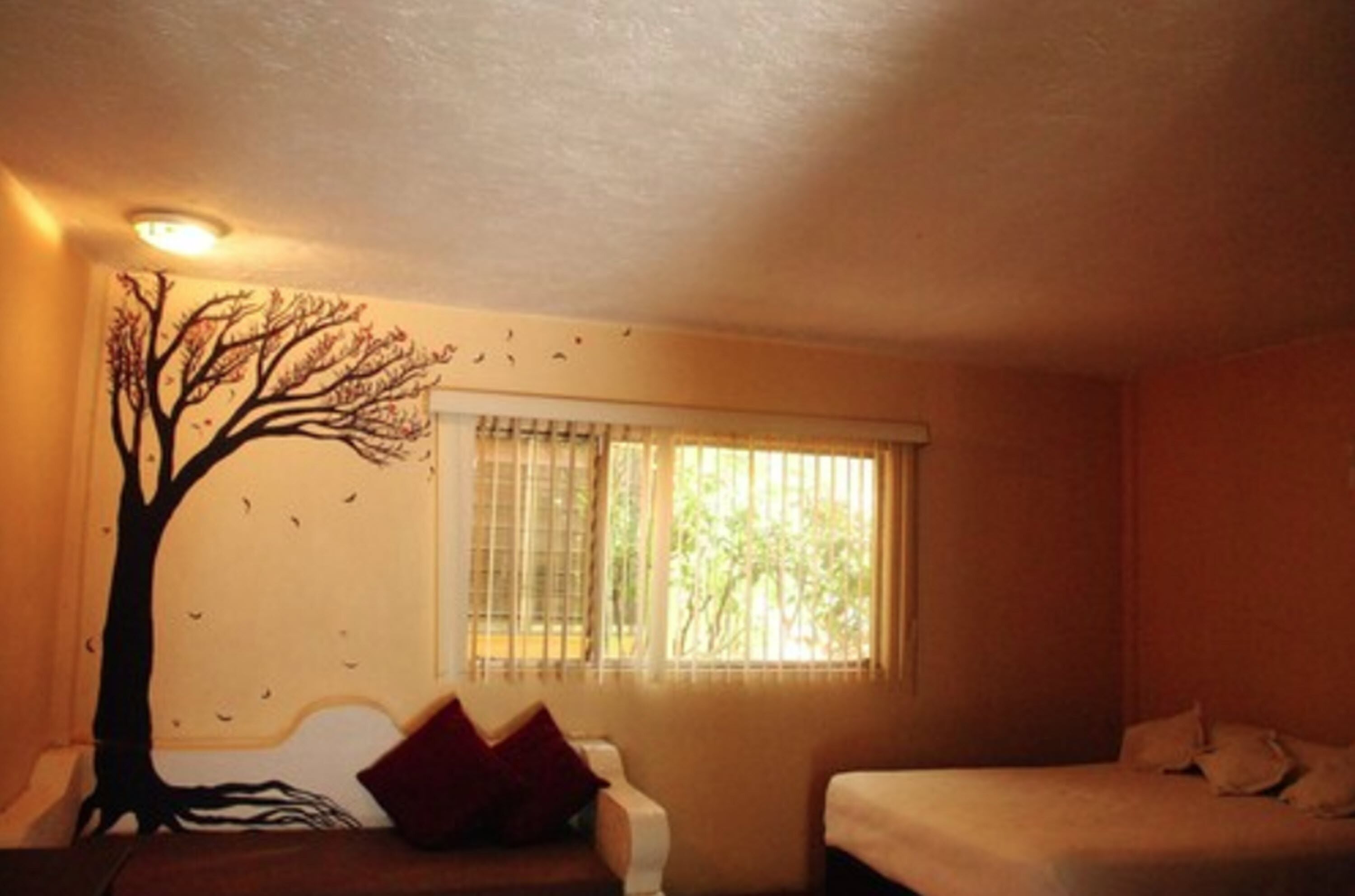 A room with a bed, a chair, a window with blinds, and a wall mural of a tree.