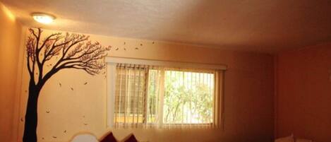 A room with a bed, a chair, a window with blinds, and a wall mural of a tree.
