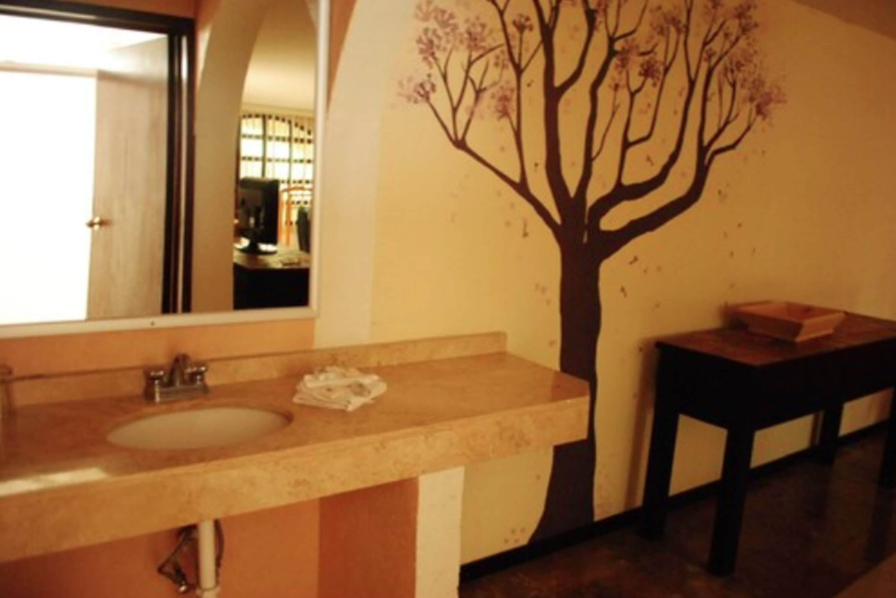 Un baño con lavamanos, espejo y un adhesivo de árbol en la pared.