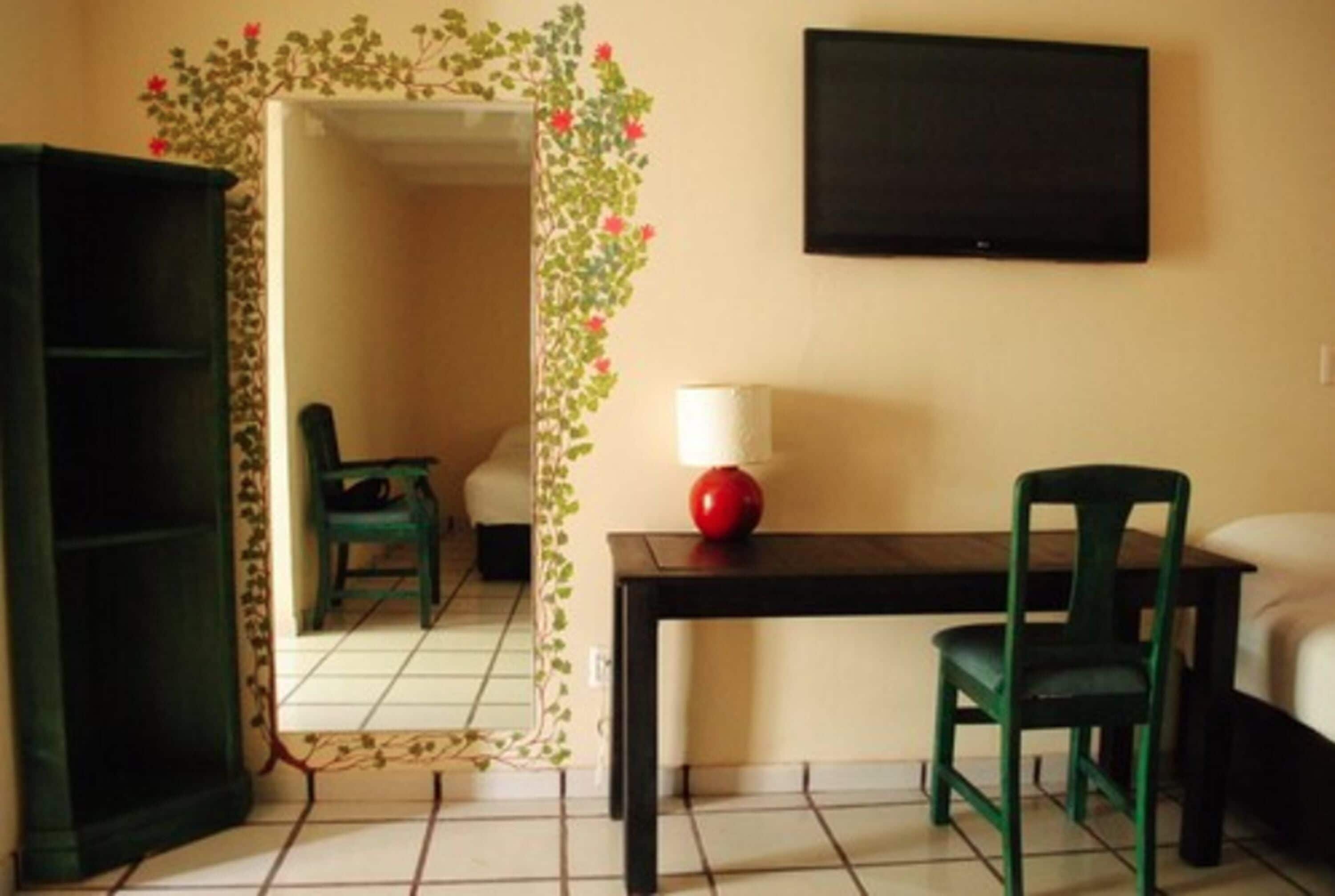 Una habitación con piso de baldosas, una silla verde, una mesa negra con una lámpara roja, un televisor negro en la pared y un espejo con marco verde.
