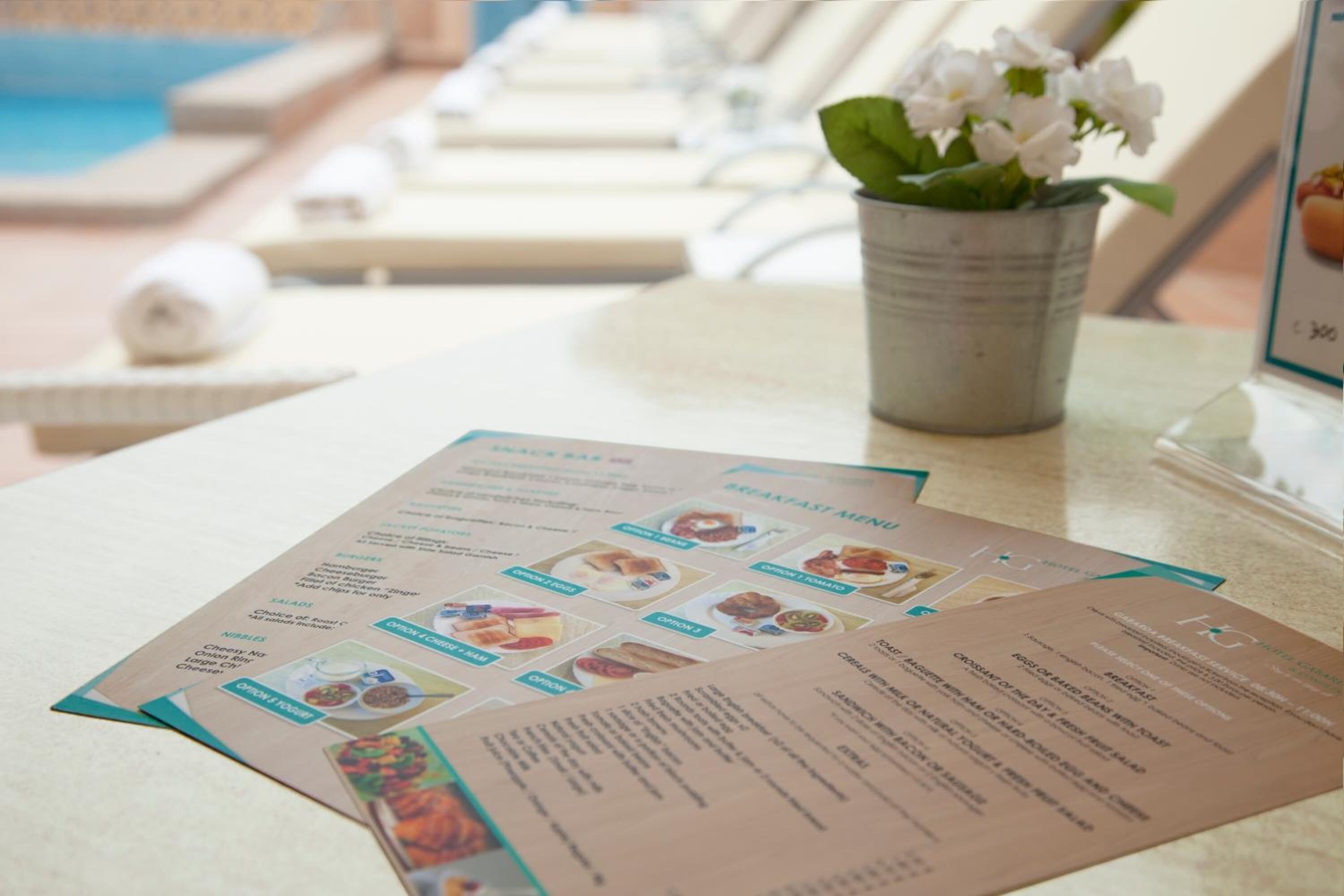 A menu with various breakfast options and a small potted plant on a table near a poolside area.