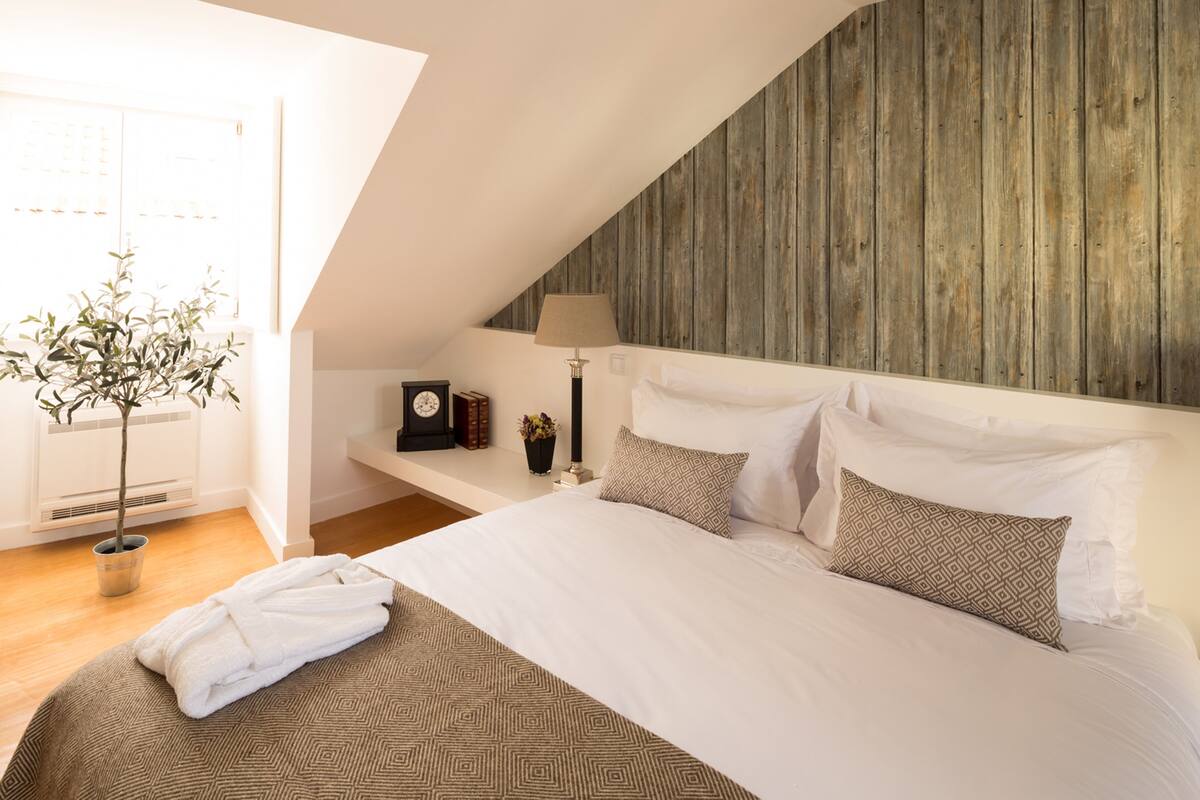 Un dormitorio con techo inclinado, cama con ropa de cama estampada, mesita de noche con lámpara, reloj y libros.