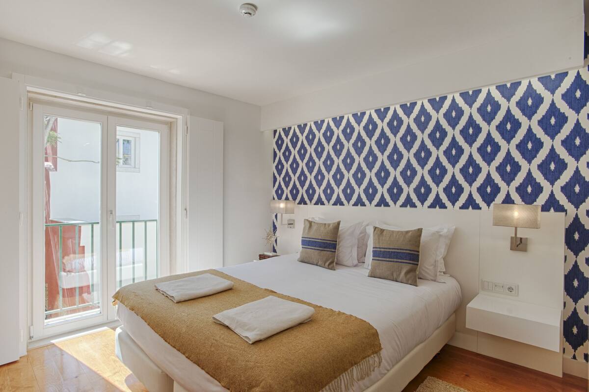 Una habitación de hotel con una cama, una mesita de noche, una lámpara de pared y un papel tapiz estampado.